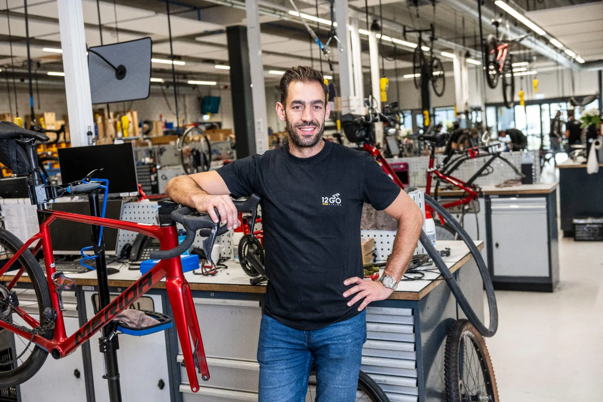 Ervaren fietstechnicus leunt lachend op een racefiets in de werkplaats van 12GO Biking